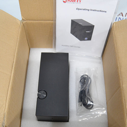 Top-down view of a 4E's USA CytoCube portable cell counter packed inside an open cardboard box with protective foam, showing the unit, an included power cable in a plastic bag, and the operating instructions manual