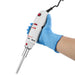 Studio product view of a gloved hand holding a 4E's USA handheld rotor-stator homogenizer against a white background, highlighting its ergonomic white body, red control switches, and attached stainless steel probe during operation