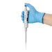 Studio product view of a 4E's USA Precipette adjustable pipette (10-100uL) held in a gloved hand against a white background, highlighting its ergonomic white body, dark blue plunger cap, digital volume window, and attached pipette tip