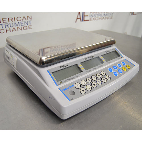 Three-quarter side-angled studio view of an Adam Equipment CBC 71a digital counting scale on a metallic surface, showing the stainless steel weighing platter, light grey casing, and front control panel with triple LCD screens and keypad