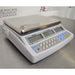 Three-quarter side-angled studio view of an Adam Equipment CBC 71a digital counting scale on a metallic surface, showing the stainless steel weighing platter, light grey casing, and front control panel with triple LCD screens and keypad