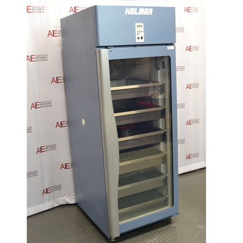 An alternative angled view of a Helmer HPR-125 pharmacy refrigerator in front of an American Instrument Exchange backdrop, displaying its closed glass door, left side panel, internal stainless steel drawers, and top blue control panel.