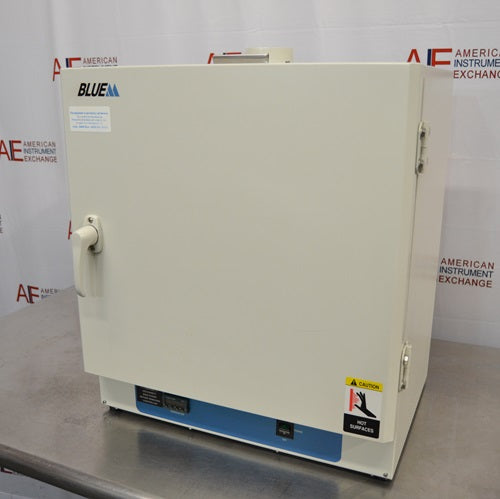 An alternative angled view of a Lindberg Blue M GO1305A laboratory oven resting on a stainless steel surface in front of an American Instrument Exchange backdrop, displaying its right side panel, door hinges, front latch handle, and bottom control panel.