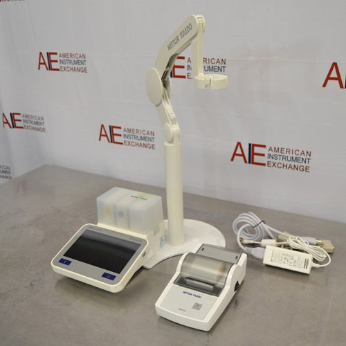 An alternative high-angled view of a Mettler Toledo SevenExcellence multiparameter meter setup on a stainless steel surface in front of an American Instrument Exchange backdrop, highlighting the touchscreen display unit, attached electrode arm, printer module, and connected cables.