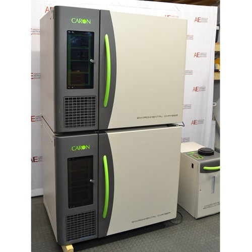 A front-facing view of the stacked Caron 6010-1 environmental chamber units, emphasizing the beige and charcoal-grey cabinet design, bright green door handles, and adjacent condensate recirculator positioned in front of an American Instrument Exchange backdrop.