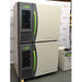 A front-facing view of the stacked Caron 6010-1 environmental chamber units, emphasizing the beige and charcoal-grey cabinet design, bright green door handles, and adjacent condensate recirculator positioned in front of an American Instrument Exchange backdrop.