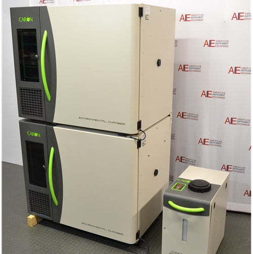 An angled view of the stacked Caron 6010-1 environmental chamber units, displaying their beige and charcoal-grey design with bright green accents, accompanied by the condensate recirculator in front of an American Instrument Exchange backdrop.