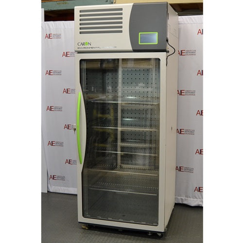 An angled view of the tall, upright Caron 7000-33-1 environmental chamber, highlighting its full-length glass door, distinctive bright green handle, and top-mounted control interface against an American Instrument Exchange backdrop.