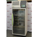 An angled view of the tall, upright Caron 7000-33-1 environmental chamber, highlighting its full-length glass door, distinctive bright green handle, and top-mounted control interface against an American Instrument Exchange backdrop.