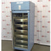 An angled view of a Helmer HPR-125 pharmacy refrigerator in front of an American Instrument Exchange backdrop, displaying its closed glass door, internal stainless steel drawers, side cabinet panel, and top blue control panel.