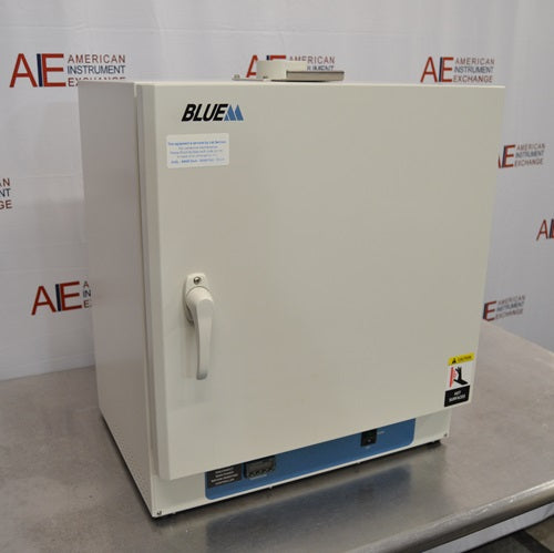 An angled view of a Lindberg Blue M GO1305A laboratory oven sitting on a stainless steel surface in front of an American Instrument Exchange backdrop, displaying its closed door, handle latch, left side panel, and bottom control panel.