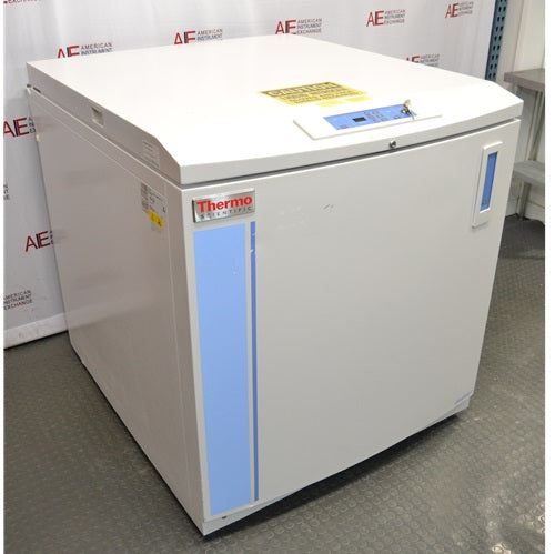 An angled view of a Thermo Scientific CryoPlus 4 cryogenic storage system resting on the floor in front of an American Instrument Exchange backdrop, showing its closed top lid, microprocessor control panel with key lock, left side casing with product label, and front face with blue vertical striping.