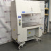 A Baker AniGARD e3 Animal Transfer Station, white in color, with a clear glass window and electronic controls on the top. It is set against a backdrop featuring the AIE Laboratory Services branding.