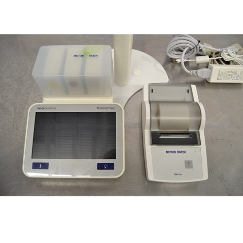 A close-up view of a Mettler Toledo SevenExcellence multiparameter meter touchscreen terminal, expansion module block, compact printer, and power supply components on a stainless steel surface.