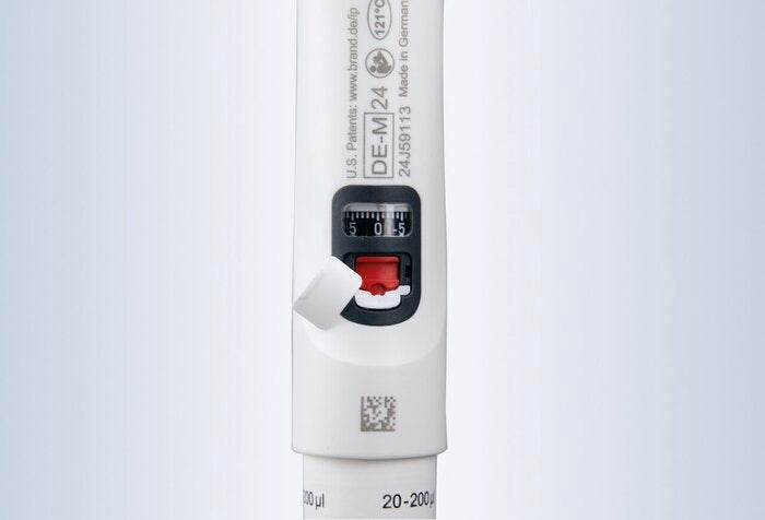 A close-up view of the handle of a white BRANDTECH Transferpette S pipette with its white protective cover flipped open, exposing the red calibration mechanism below the digital volume display window.