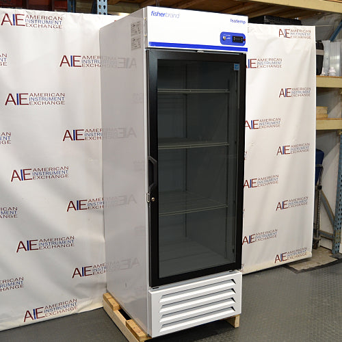 Straight-on studio view of a Fisherbrand Isotemp FBG25RPGA upright laboratory refrigerator standing on a wooden pallet against a branded backdrop, featuring a full-length black-framed glass door, top-mounted digital display console, and louvered white base