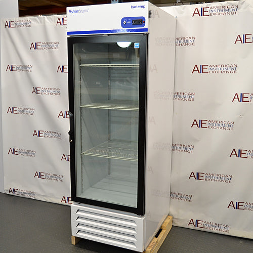 Straight-on studio view of a Fisher Isotemp GTFBG25RPGA upright laboratory refrigerator standing on a wooden pallet against a branded backdrop, featuring a full-length glass door, top-mounted digital controller, Fisherbrand logo, and lower ventilation louvers