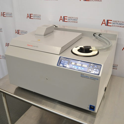 A front-facing angled view of a Thermo Savant SpeedVac SPD2030 vacuum concentrator system on a stainless steel lab table, showing its integrated control panel, vacuum tubing, and top cover housing.