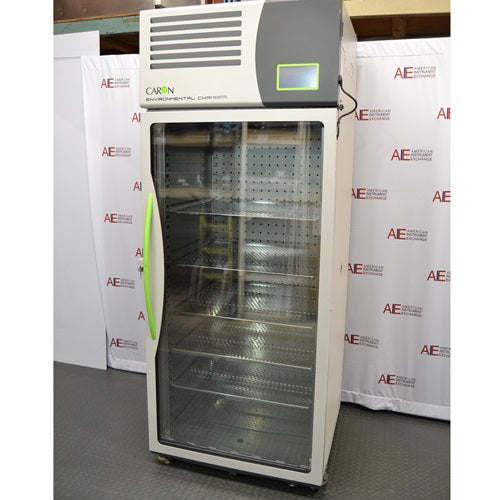 A front-facing view of the tall, upright Caron 7000-33-1 environmental chamber, highlighting its full-length glass door, distinctive bright green handle, and top-mounted control interface against an American Instrument Exchange backdrop.