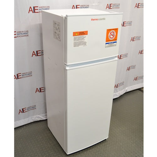 A front-left angled view of a white Thermo Scientific flammable material combination refrigerator/freezer on a lab floor, showing safety warning labels and serial specifications stickers on the upper freezer section against an American Instrument Exchange backdrop.