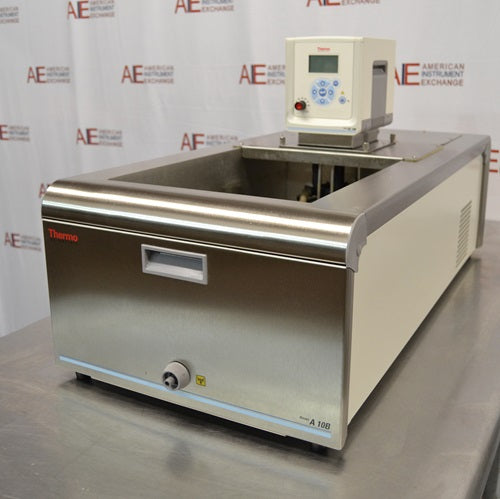 An angled front-side view of a Thermo Scientific A10B refrigerated bath circulator sitting on a stainless steel surface in front of an American Instrument Exchange backdrop, displaying the stainless steel front panel with drain port, open top bath area, mounted digital control head with display screen, and side ventilation louvers.