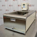 An angled front-side view of a Thermo Scientific A10B refrigerated bath circulator sitting on a stainless steel surface in front of an American Instrument Exchange backdrop, displaying the stainless steel front panel with drain port, open top bath area, mounted digital control head with display screen, and side ventilation louvers.