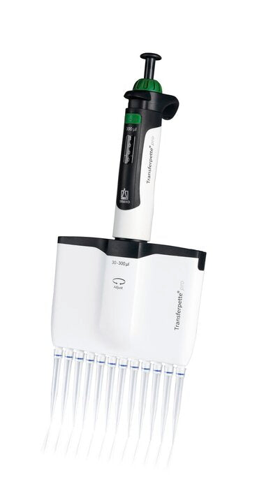 A front view of an upright BRANDTECH Transferpette S 12-channel adjustable pipette featuring a white body with green accents, a digital volume display window, top plunger button, and twelve clear pipette tips attached at the base.