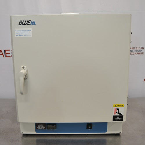 A front-facing view of a Lindberg Blue M GO1305A laboratory oven sitting on a stainless steel surface, featuring a solid front door with a latch handle, Blue M branding, hot surface warning label, and bottom control panel.