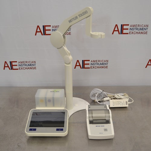 A front-facing view of a Mettler Toledo SevenExcellence multiparameter meter setup on a stainless steel surface in front of an American Instrument Exchange backdrop, featuring a touchscreen main unit, attached electrode arm, printer module, and power supply cables.