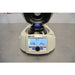 A front view of an open Oxford Lab Products Benchmate C12V microcentrifuge showing the internal rotor, central blue locking knob, control dial, and illuminated digital display screen with speed and timer settings against an American Instrument Exchange backdrop.