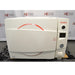 A front-facing view of a Tuttnauer 3870ELP-B/L-D benchtop autoclave sitting on a pallet, featuring a white main door with a red circular accents, Tuttnauer logo, digital display panel, pressure gauge, and built-in paper printer.
