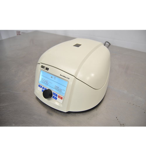 An elevated front-left view of a closed Oxford Lab Products Benchmate C12V microcentrifuge sitting on a stainless steel surface, highlighting the top lid, power cord, and front control panel with illuminated display.