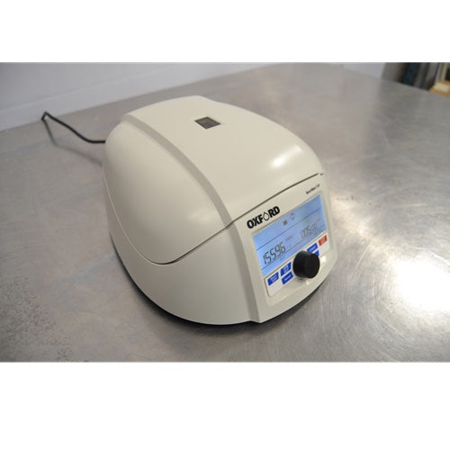 An elevated front-left view of a closed Oxford Lab Products Benchmate C12V microcentrifuge sitting on a stainless steel surface, highlighting the top lid, power cord, and front control panel with illuminated display.