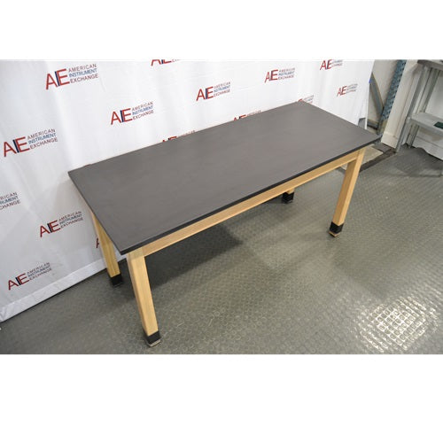 An elevated front-left view of a wooden laboratory bench with a black epoxy resin tabletop supported by four light wood legs with protective black base caps against an American Instrument Exchange backdrop.