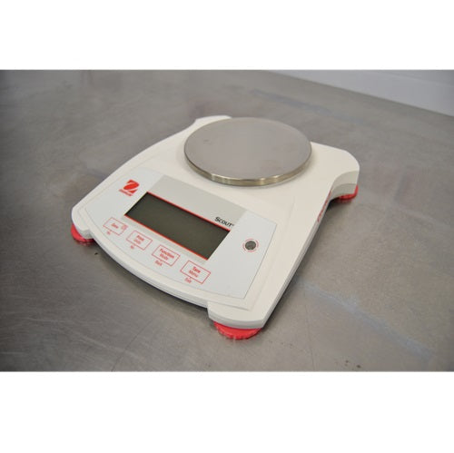 An elevated front-left view of a white Ohaus SPX222 Scout portable balance resting on a metal surface, featuring a round stainless steel weighing pan, front display screen, control buttons, and red support feet.