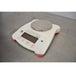 An elevated front-left view of a white Ohaus SPX222 Scout portable balance resting on a metal surface, featuring a round stainless steel weighing pan, front display screen, control buttons, and red support feet.