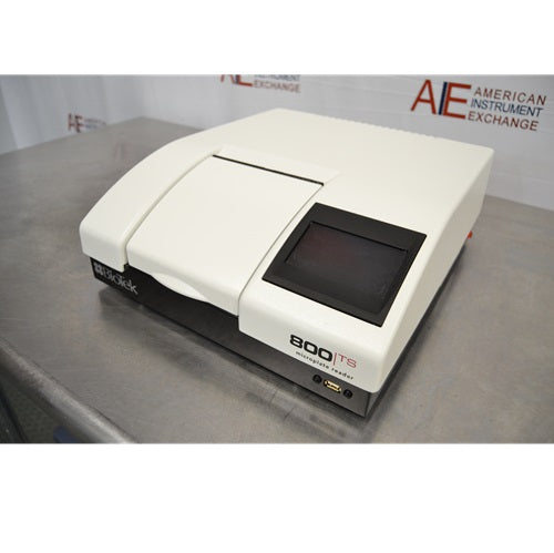An elevated front-right view of a white and black BioTek 800 TS absorbance microplate reader sitting on a stainless steel surface, featuring the top compartment cover, front touchscreen display, and lower USB port against an American Instrument Exchange backdrop.