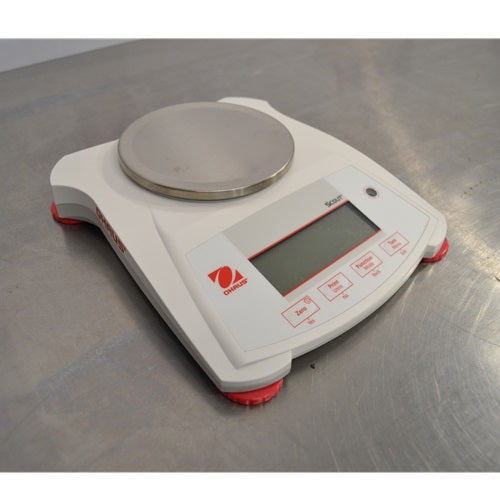 An elevated front-right view of a white Ohaus SPX222 Scout portable balance on a stainless steel tabletop, displaying the circular metal weighing plate, red side branding, front control panel, and adjustable red feet.