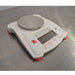 An elevated front-right view of a white Ohaus SPX222 Scout portable balance on a stainless steel tabletop, displaying the circular metal weighing plate, red side branding, front control panel, and adjustable red feet.