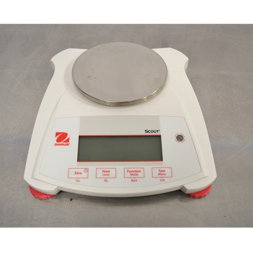 An elevated front view of a white Ohaus SPX222 Scout portable balance sitting on a metal surface, featuring a round stainless steel weighing pan, digital display screen, red leveling feet, and front control buttons.