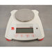 An elevated front view of a white Ohaus SPX222 Scout portable balance sitting on a metal surface, featuring a round stainless steel weighing pan, digital display screen, red leveling feet, and front control buttons.