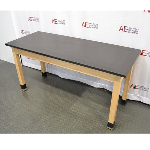 An elevated side-angled view of a wooden laboratory bench featuring a smooth black epoxy resin top supported by light wood legs with protective black base boots against an American Instrument Exchange backdrop.