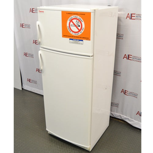 An elevated side-angle view of a white Thermo Scientific 10FCEETSA flammable material combination refrigerator/freezer on a lab floor, showing the top freezer door with a safety warning sign and the lower refrigerator door against an American Instrument Exchange backdrop.