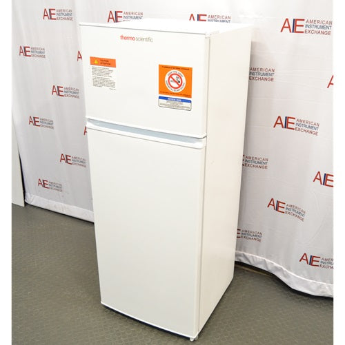 An elevated front-angled view of a white Thermo Scientific flammable material combination refrigerator/freezer, showing safety warning labels on the top freezer door and the main refrigerator compartment door against an American Instrument Exchange backdrop.