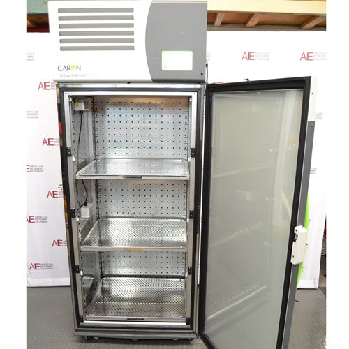 The interior of an open Caron 7401-33-22 reach-in CO₂ incubator, featuring adjustable stainless steel shelves, perforated side walls, and an inner glass door, set against an American Instrument Exchange backdrop.