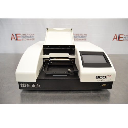 A front view of a white and black BioTek 800 TS absorbance microplate reader with its top cover door open, revealing the internal microplate carrier tray, featuring a touchscreen display on the right and a USB port on the lower front panel against an American Instrument Exchange backdrop.