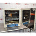 An open-door view of a Lindberg Blue M GO1305A laboratory oven on a stainless steel surface, revealing the interior stainless steel chamber, perforated shelving, door gasket, and reflective inner door panel.