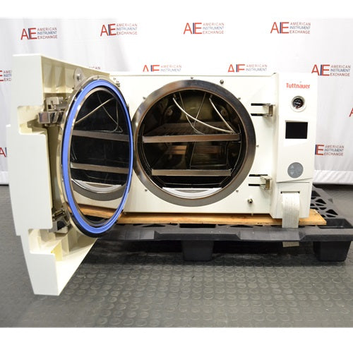 An open-door view of a Tuttnauer 3870ELP-B/L-D benchtop autoclave resting on a pallet in front of an American Instrument Exchange backdrop, displaying its open front door with blue seal gasket, interior cylindrical stainless steel sterilization chamber, internal rack shelving, and right side control interface.