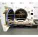An open-door view of a Tuttnauer 3870ELP-B/L-D benchtop autoclave resting on a pallet in front of an American Instrument Exchange backdrop, displaying its open front door with blue seal gasket, interior cylindrical stainless steel sterilization chamber, internal rack shelving, and right side control interface.