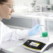 A laboratory technician wearing safety glasses and protective gloves uses a micro-spatula to add powder into a glass beaker of green liquid inside the draft shield chamber of a Sartorius Secura 120 analytical balance